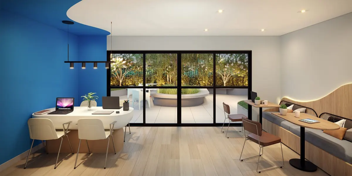 O coworking com uma parede azul, mesas de trabalho e a vista do jardim pelo vidro.