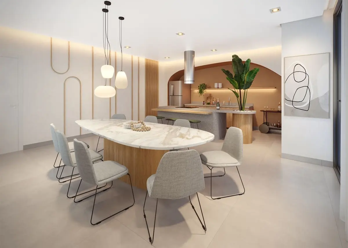O espaço gourmet com uma mesa oval de mármore sob pendentes e a cozinha ao fundo.